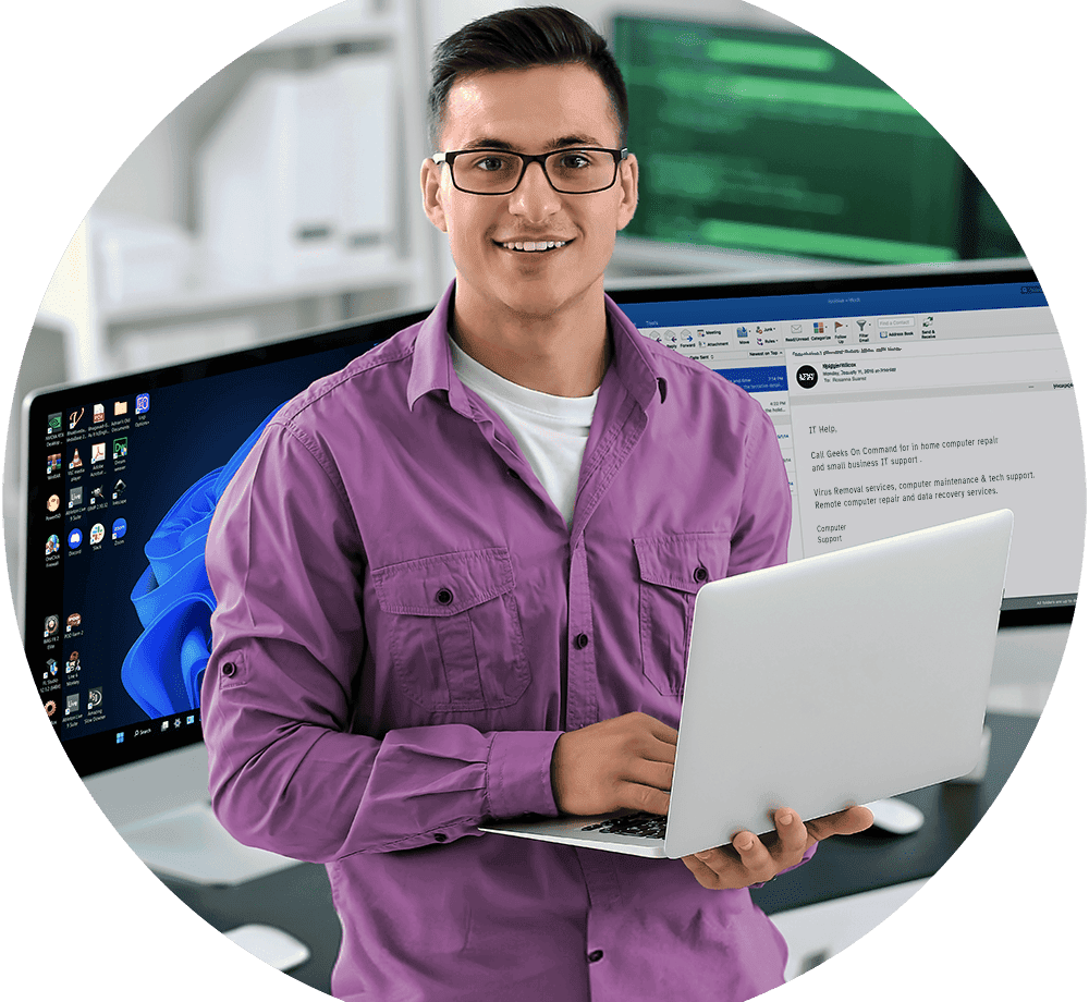 Smiling, confident young male IT professional in glasses and a purple button-down providing computer support on a laptop in a home office, with dual monitors showing a tech support email about Geeks On Command's in-home computer repair, remote IT support, and data recovery services.