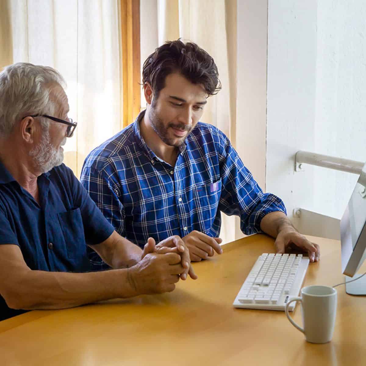 An older man receiving computer help at home from a younger man, both seated at a desk with a desktop computer.